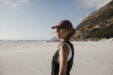 Südafrika, Westkap, Noordhoek Beach, lächelnde junge Frau mit Basecap am Strand stehend - LHPF00402