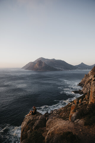 Südafrika, Westkap, Frau sitzt auf einem Felsen und telefoniert, gesehen vom Chapman's Peak Drive, lizenzfreies Stockfoto