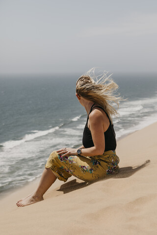 Namibia, Walvis Bay, Namib-Naukluft-Nationalpark, Sandwich Harbour, Frau sitzt in Dünenlandschaft am Meer, lizenzfreies Stockfoto