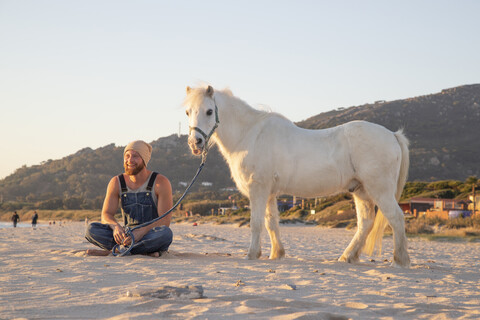Spanien, Tarifa, glücklicher Mann mit Pony am Strand sitzend, lizenzfreies Stockfoto