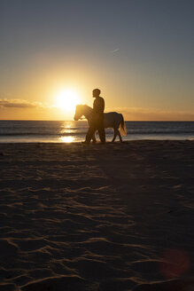 Spanien, Tarifa, Mann geht mit Pony am Strand bei Sonnenuntergang spazieren - KBF00482