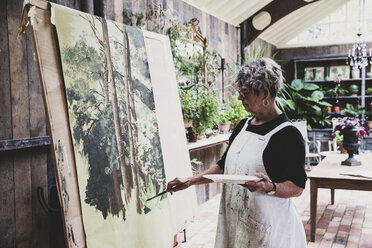 Ältere Frau mit Brille, schwarzem Oberteil und weißer Schürze steht im Atelier und arbeitet an einem Bild von Bäumen im Wald. - MINF10215