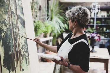 Ältere Frau mit Brille, schwarzem Oberteil und weißer Schürze steht im Atelier und arbeitet an einem Bild von Bäumen im Wald. - MINF10216