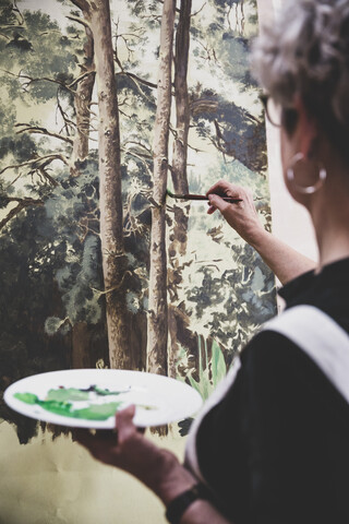 Ältere Frau mit Brille, schwarzem Oberteil und weißer Schürze steht im Atelier und arbeitet an einem Bild von Bäumen im Wald., lizenzfreies Stockfoto