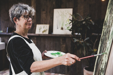Ältere Frau mit Brille, schwarzem Oberteil und weißer Schürze steht im Atelier und arbeitet an einem Bild von Bäumen im Wald. - MINF10219