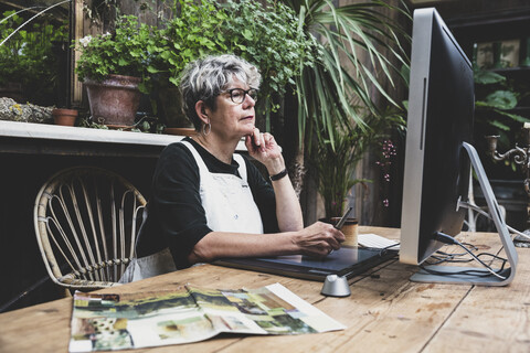 Ältere Frau mit Brille, schwarzem Oberteil und weißer Schürze sitzt an einem Holztisch und arbeitet an einem Desktop-Computer., lizenzfreies Stockfoto