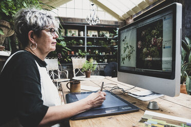 Ältere Frau mit Brille, schwarzem Oberteil und weißer Schürze sitzt an einem Holztisch und arbeitet an einem Desktop-Computer. - MINF10224