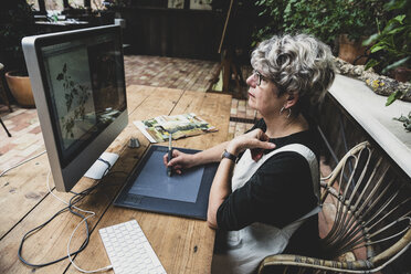 Ältere Frau mit Brille, schwarzem Oberteil und weißer Schürze sitzt an einem Holztisch und arbeitet an einem Desktop-Computer. - MINF10225