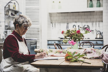 Senior woman wearing glasses, red dress and white apron sitting at table, working on pencil drawing of orange Dahlia. - MINF10257