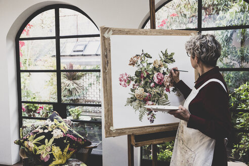 Ältere Frau mit Brille, rotem Kleid und weißer Schürze steht im Atelier und arbeitet an einem Bild mit rosa Teerosen, Blättern, Beeren und anderen Blumen. - MINF10260