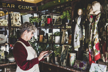 Ältere Frau mit Brille, rotem Kleid und weißer Schürze steht in einem Geschäft für Innenarchitektur und hält eine digitale Tabelle. - MINF10267
