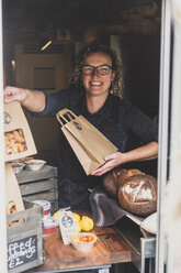 Eine lächelnde Frau mit Brille steht in einer Bäckerei und hält eine braune Papiertüte in der Hand. - MINF10304