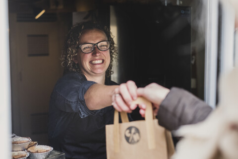 Lächelnde Frau mit Brille, die eine braune Papiertüte durch das Fenster einer Bäckerei reicht., lizenzfreies Stockfoto