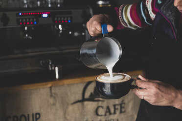 Close up of barista pouring milk from jug into cappuccino mug, creating foam pattern. - MINF10329