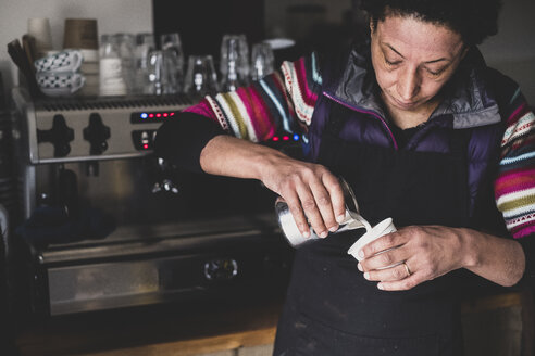Frau hinter Espressomaschine gießt heiße Milch in Pappbecher. - MINF10331