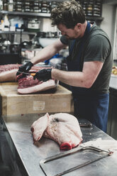 Männlicher Metzger mit Schürze und schwarzen Gummihandschuhen beim Schneiden von Schweinerippchen auf einem Fleischerblock. - MINF10366