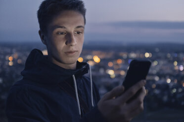 Porträt eines jungen Mannes mit Blick auf ein leuchtendes Smartphone am Abend - GCF00239