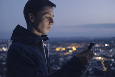 Junger Mann schaut abends auf sein glänzendes Smartphone - GCF00240