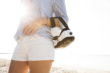 Mann mit Virtual-Reality-Brille auf dem Arm, am Strand gegen die Sonne - HMEF00199