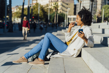 Beautiful woman sitting on stairs in the city, using smartphone, listening music with headphones - BOYF01332