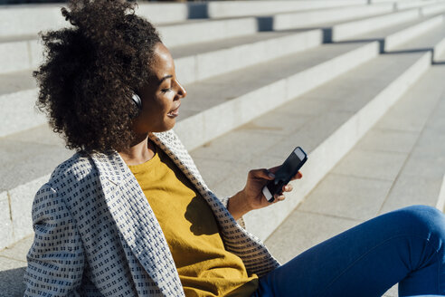 Schöne Frau, die auf einer Treppe in der Stadt sitzt, ein Smartphone benutzt und mit Kopfhörern Musik hört - BOYF01334