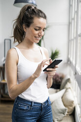 Smiling woman checking cell phone at the window at home - GIOF05640