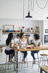 Three happy women sitting at kitchen table at home socializing - GIOF05645