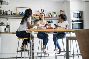 Drei glückliche Frauen sitzen am Küchentisch zu Hause und unterhalten sich - GIOF05646