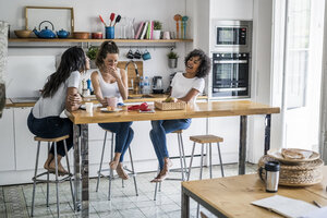 Drei glückliche Frauen sitzen am Küchentisch zu Hause und unterhalten sich - GIOF05647