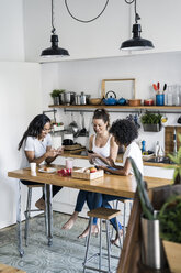 Drei glückliche Frauen sitzen zu Hause am Küchentisch und benutzen Handys - GIOF05648
