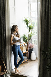 Woman with cup of coffee and headphones standing in doorframe at home - GIOF05666