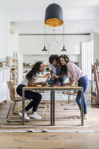 Drei glückliche Frauen mit digitalen Geräten auf dem Tisch zu Hause, lizenzfreies Stockfoto