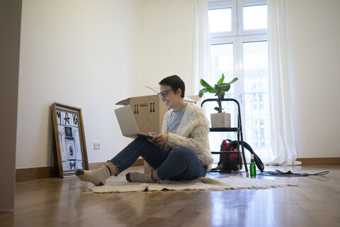 Junge Frau sitzt in ihrer neuen Wohnung auf dem Boden und benutzt einen Laptop, lizenzfreies Stockfoto