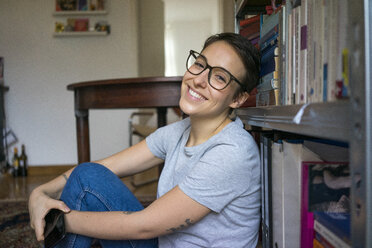 Glückliche junge Frau, die zu Hause sitzt und sich auf ein Bücherregal stützt - MOEF01943