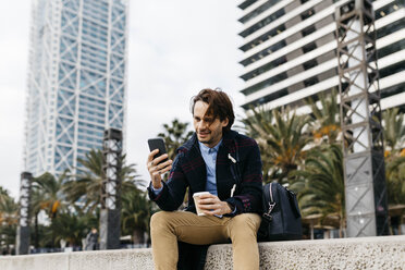 Spanien, Barcelona, lächelnder Mann mit Kaffee zum Mitnehmen und Mobiltelefon in der Stadt - JRFF02501