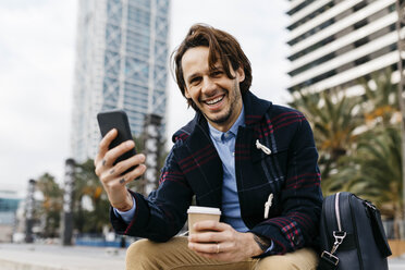 Spanien, Barcelona, glücklicher Mann mit Kaffee zum Mitnehmen und Mobiltelefon in der Stadt - JRFF02504