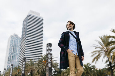 Spain, Barcelona, smiling man walking in the city with two skyscrapers in the background - JRFF02506