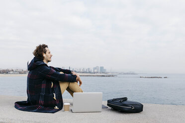 Spanien, Barcelona, Mann mit Laptop, Aktentasche und Kopfhörern am Meer sitzend - JRFF02516