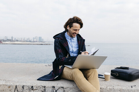 Spanien, Barcelona, lächelnder Mann sitzt am Meer und arbeitet mit Laptop und Notebook, lizenzfreies Stockfoto