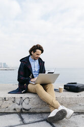 Spanien, Barcelona, Mann sitzt am Meer und arbeitet mit Laptop und Notizbuch - JRFF02519