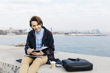 Spain, Barcelona, portrait of man sitting at the sea taking notes - JRFF02524