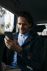 Smiling man sitting on back seat of a car using cell phone - JRFF02527