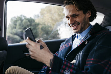 Smiling man sitting on back seat of a car using cell phone - JRFF02529