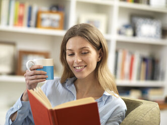Lächelnde junge Frau mit einem heißen Getränk, die sich zu Hause beim Lesen eines Buches entspannt - ABRF00283