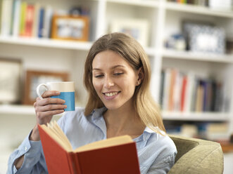 Smiling young woman with a hot drink relaxing at home reading a book - ABRF00283