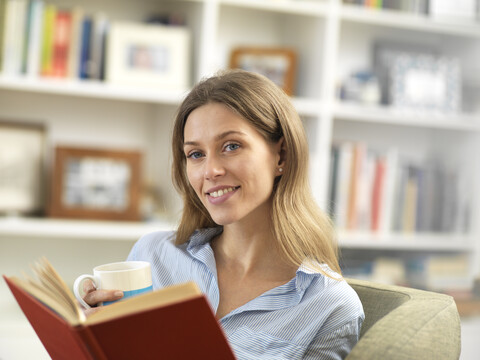 Lächelnde junge Frau entspannt sich zu Hause mit einem heißen Getränk und einem Buch, lizenzfreies Stockfoto