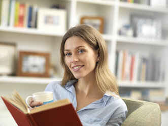 Lächelnde junge Frau entspannt sich zu Hause mit einem heißen Getränk und einem Buch - ABRF00284