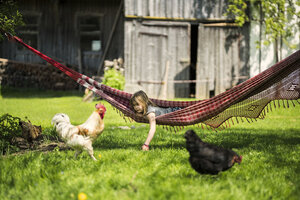 Mädchen entspannt in Hängematte im Garten eines Bauernhofs mit Huhn im Vordergrund - SBOF01699