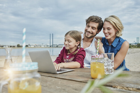 Germany, Duesseldorf, happy family with daughter using laptop on wooden table at Rhine riverbank - RORF01710