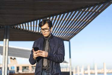 Portrait of fashionable young businessman wearing glasses and tie looking at mobile phone - AFVF02314